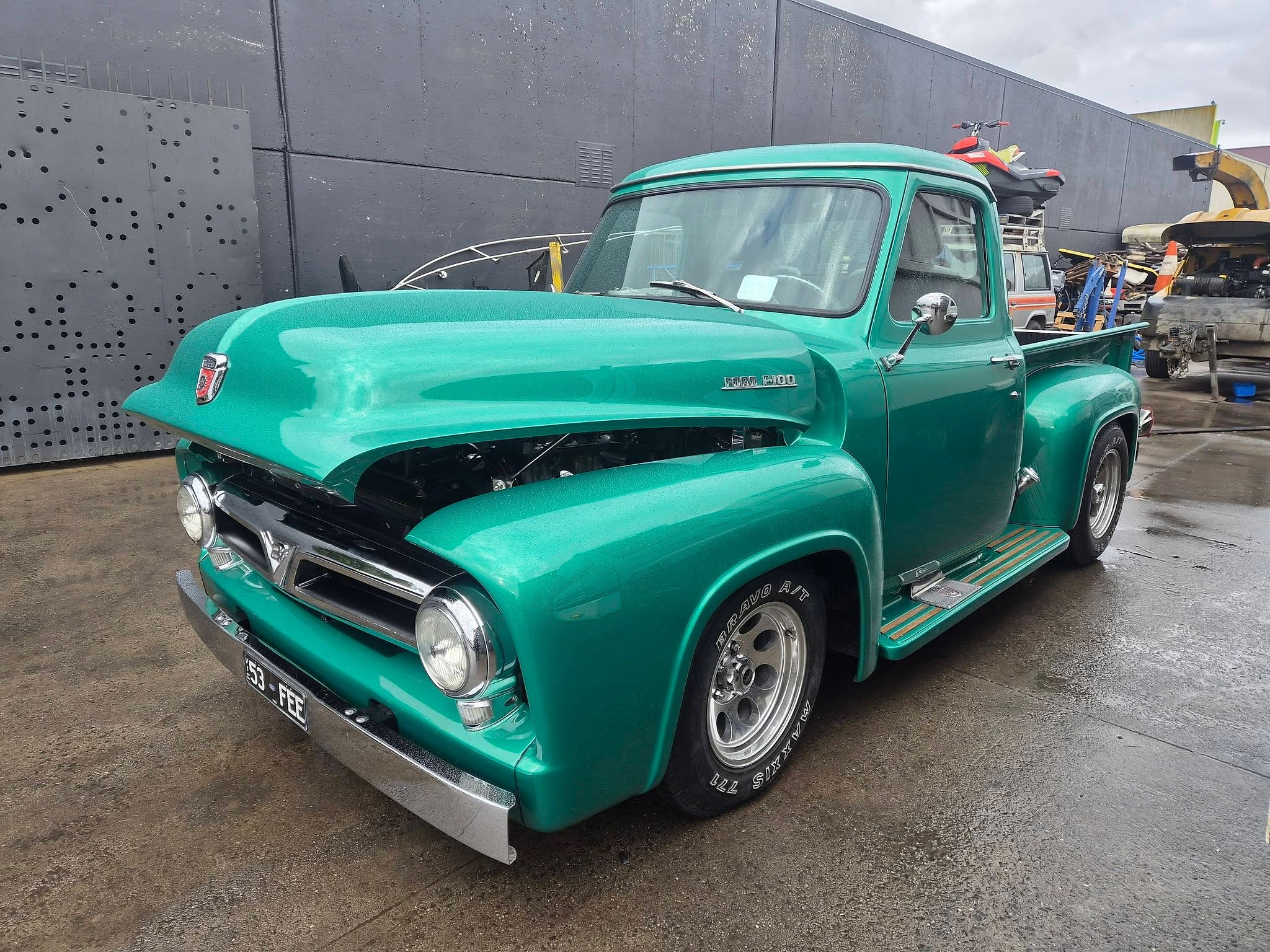 A Ford F100 pickup in deep teal, front three quarter, on the concrete outside the shop