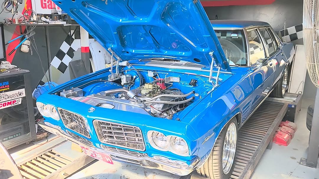 The front end of a blue Falcon on the workshop floor with the bonnet raised and the front panels off