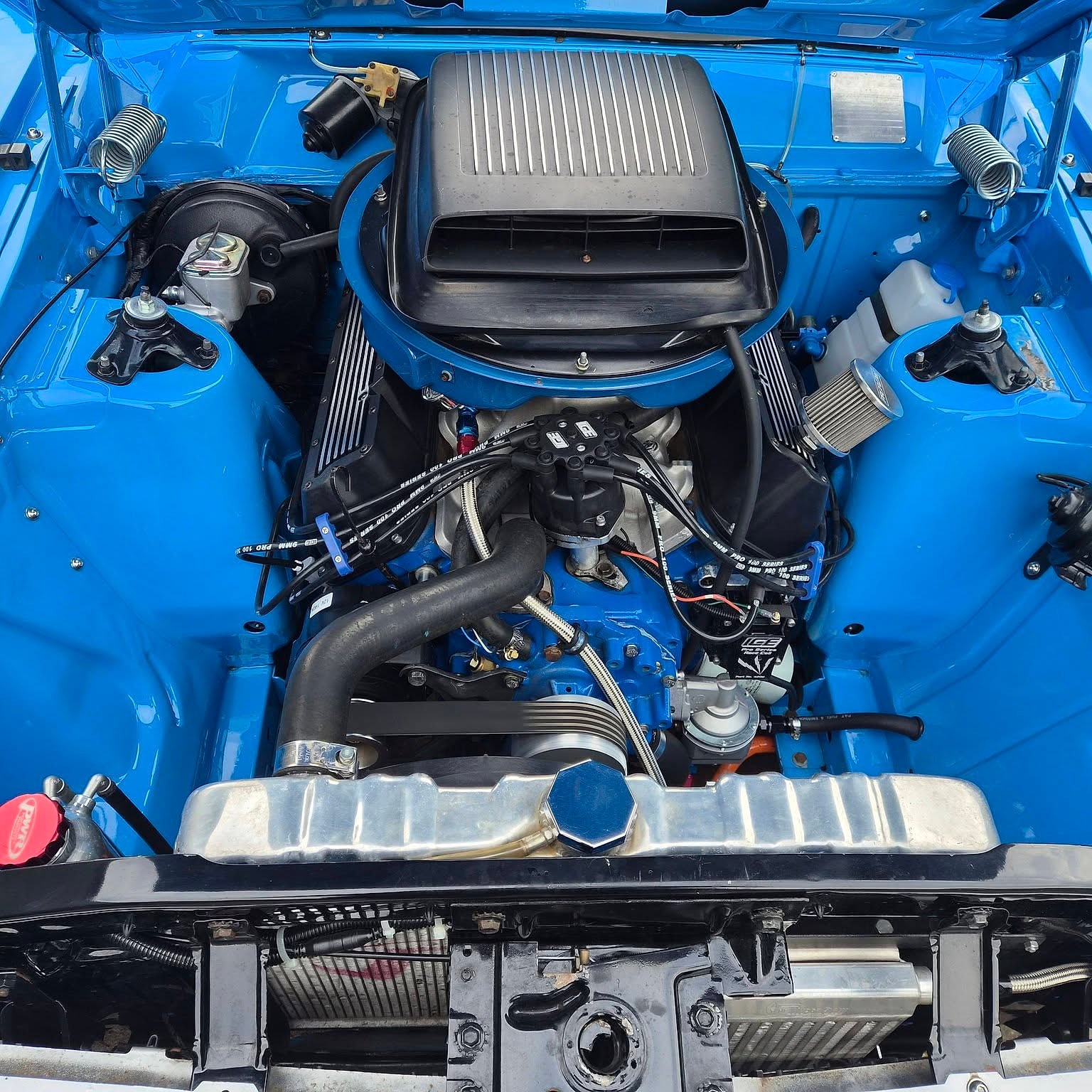A blown V8 in a bright blue engine bay, the supercharger and scoop standing up through the bonnet opening