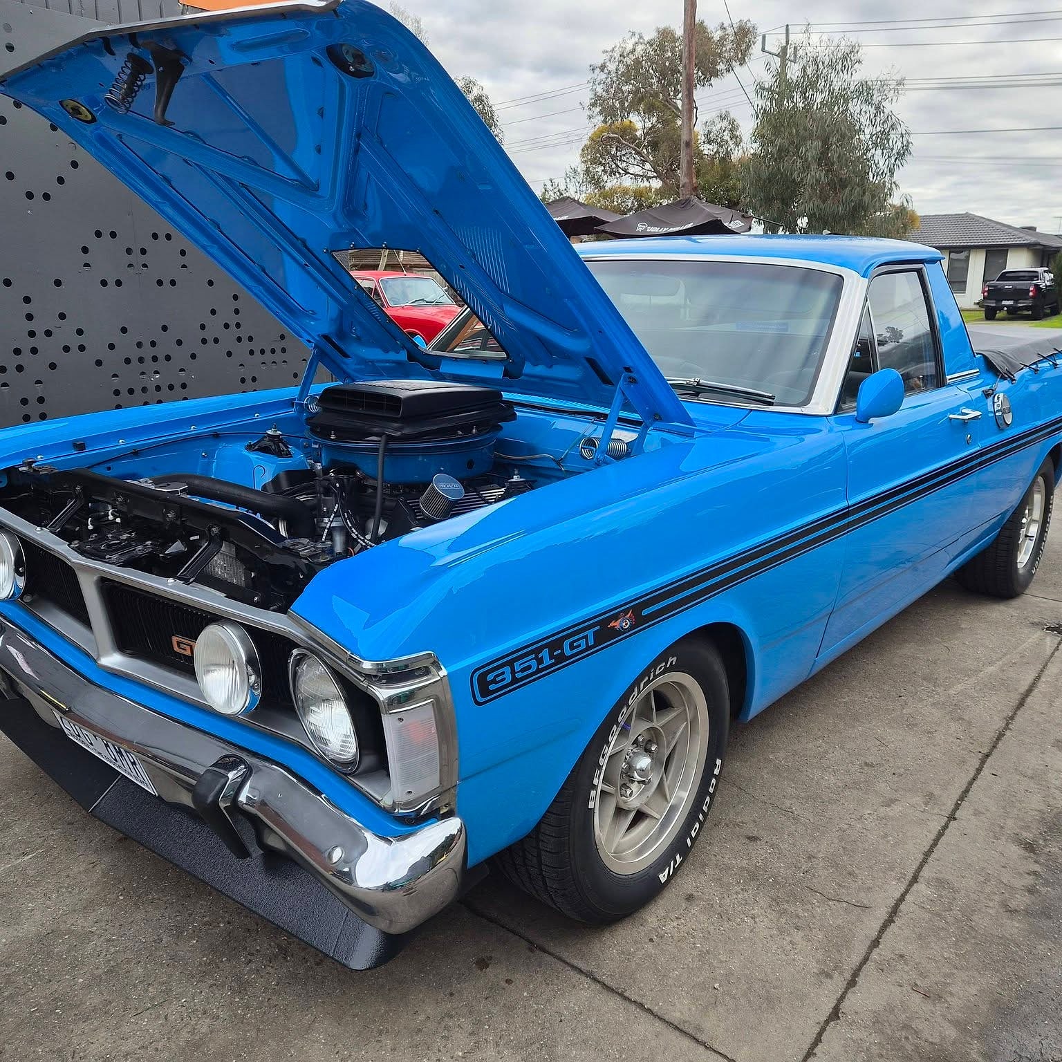 A Ford Falcon GT ute in bright blue with the bonnet up, showing a blown V8, parked outside the workshop