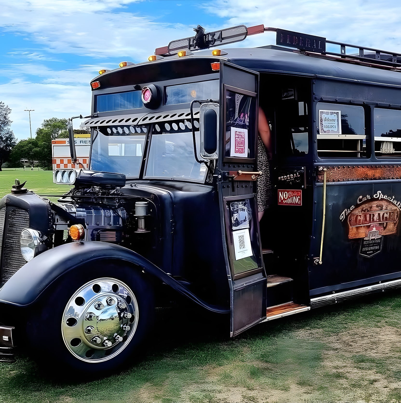 The Car Specialist Hotrod Bus, matt black with a blown engine standing out of the bonnet and the Garage signwriting on its side, parked on the grass at a show