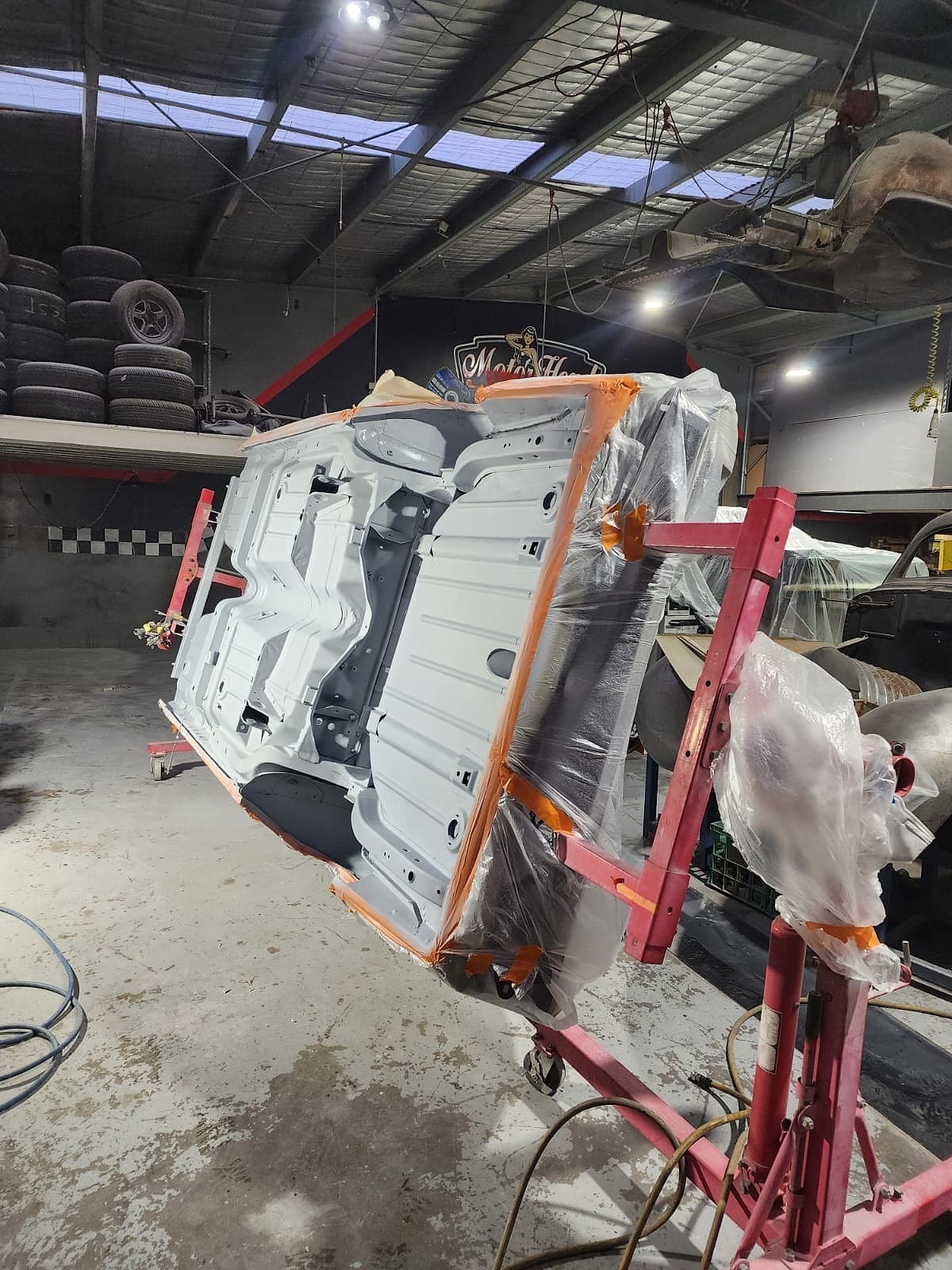 A car body shell turned on its side on a rotisserie in the workshop, the underside in grey primer and the edges masked in plastic and orange tape