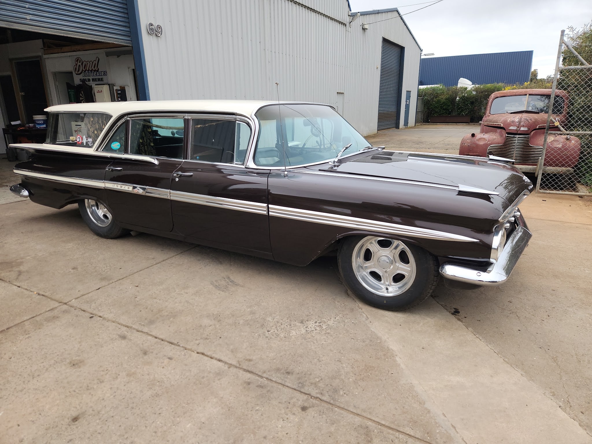 A long American wagon in maroon over cream parked outside the workshop, with a rusted 1940s coupe behind the fence